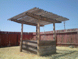A low resolution, dithered photograph of a wooden planter structure in a garden. Sun is out, and there's an orange fence in the background.